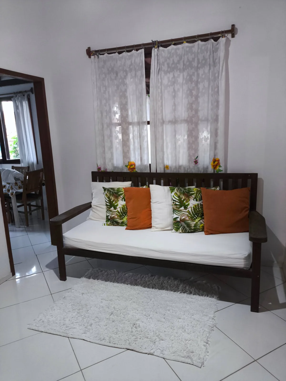 Sala de estar da casa de temporada em Trancoso BA, com sofá de madeira, almofadas laranja e cortinas de renda